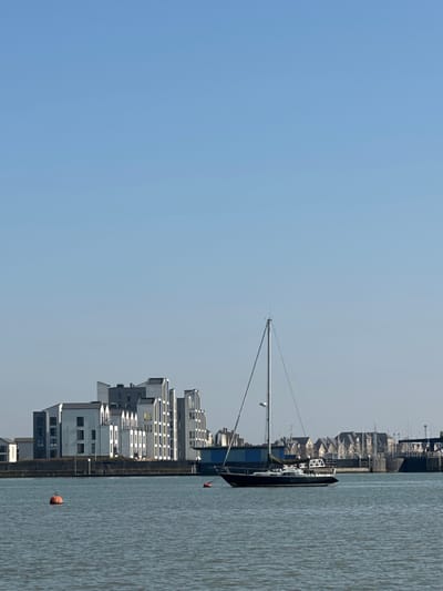 A clear blue sky occupies the upper frame, above a calm body of water. In the midground, a dark-hulled sailboat with a prominent silver mast is moored, positioned slightly right of the image center. No sails are deployed, and no individuals are visible on board. Two orange buoys float in the water: one in the mid-left foreground and a smaller one further back to the right of the first. The background features a modern architectural development along the waterfront in Rochester, United Kingdom. These buildings are predominantly white and grey, exhibiting multi-story designs with angular, geometric facades and varied rooflines. Some windows appear illuminated. To the far right, beyond the sailboat, an array of residential buildings with dark roofs and a large structural element, consistent with a bridge or lock system on the River Medway, are visible. The scene is devoid of human presence, depicting a tranquil waterfront under bright, sunny conditions.