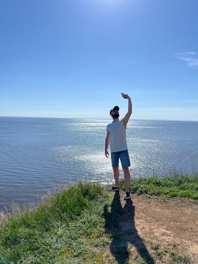 A full body shot of a person on the shore. The person is wearing a white t-shirt, blue shorts, and a black cap. The person has one arm raised, and the other arm is at their side. The water is sparkling in the sunlight. The sky is blue and sunny. The photo was taken outside near Шуран, Russia.