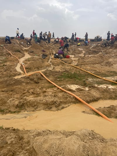 The image depicts an outdoor scene in Maiyanga, Nigeria, showing an area of dirt and mud where a large group of people is gathered. The individuals appear to be working or digging in the dirt. Several orange hoses are visible across the terrain, one leading to a narrow channel of muddy water. The sky is overcast.