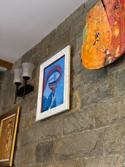 The image shows a stone wall in Кирково, Bulgaria, with several framed artworks hanging on it. On the upper left, there is a large, irregularly shaped artwork in shades of orange and brown. Below it, a rectangular painting with a white frame depicts a woman in a blue and red headscarf. Below this painting, there is a wall sconce with two white cylindrical lights mounted on a black bracket. To the lower right, a portion of another framed artwork is visible, featuring golden hues. The scene is indoors, possibly in a home or gallery.