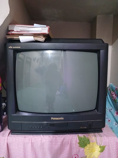 A Panasonic brand CRT television, black in color, rests on a pink and white checkered cloth draped over a surface. The television has a screen that reflects a dimly lit room, showing a person's silhouette, and a stack of books with red and brown covers is placed on top of it. The television's front panel displays buttons and the "Panasonic" logo, along with "4 SYSTEM" text. The scene appears to be indoors, possibly in a room with muted lighting, and the background wall is a pale color. The location of Geudonggeudong, Indonesia, is mentioned in the prompt but is not visually evident in the image itself.