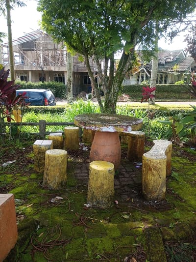 A quaint outdoor setting features a round stone table surrounded by matching cylindrical stools. Moss covers the brick ground and the bases of the seating, lending a weathered charm. The scene is partially shaded by a large tree, with buildings visible in the background – one under construction. A dark vehicle sits parked nearby. The overall aesthetic evokes a tranquil, slightly overgrown garden space.