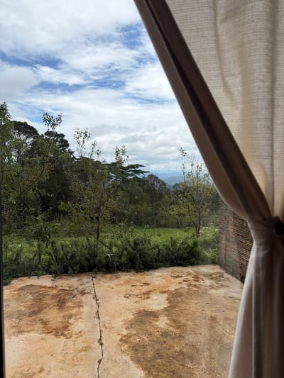 The image captures a view from an indoor space looking outwards through a partially drawn curtain. The scene depicts a natural landscape with lush green vegetation, trees, and distant mountains under a cloudy sky. The foreground shows a concrete patio with irregular brown stains. To the right, a portion of a brick wall is visible, and a cream-colored curtain hangs on the right side of the frame, obscuring part of the view. The environment suggests a location with a tropical or subtropical climate, possibly a resort or a private residence situated in a mountainous area. The overall impression is one of serene natural beauty and a connection to the outdoors. The image does not contain any people or explicit actions, focusing solely on the environment and architectural elements.