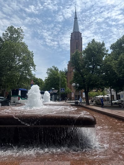 This media file shows a street scene in Bonn, Germany, with a focus on a water fountain in the foreground. The fountain has two spouts spraying water upwards, creating a foamy effect. The fountain structure appears to be made of stone with a reddish-brown hue.

In the background, a tall, narrow church tower with a pointed steeple dominates the skyline. The tower is made of brick and has a cross at its very peak. Lush green trees line the street, their leaves providing shade. Beyond the trees, buildings can be seen, some appearing to be shops or residential structures.

Several people are visible in the scene, though not clearly defined. Some appear to be walking or standing on the sidewalk, while others are seated at tables, possibly at an outdoor cafe. A bus with the text "BISCHOFF" is parked on the street, along with a car. Traffic signs are also visible, including a blue square sign with a white pedestrian symbol and a yellow triangular sign with a red border and a black symbol indicating a pedestrian crossing.

The sky is partly cloudy, with white, wispy clouds scattered across a pale blue expanse. The lighting suggests it is daytime, likely midday or early afternoon. The overall atmosphere appears to be relaxed and leisurely, typical of a pleasant day in a European city. The presence of trees, people outdoors, and a fountain suggests an urban public space.