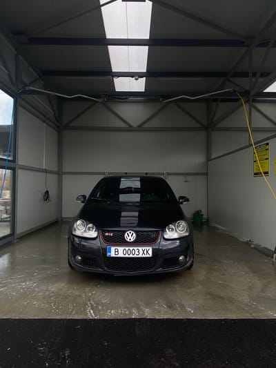 The image captures a black Volkswagen Golf GTI parked inside a self-service car wash bay. The car is centered in the frame, facing forward, showcasing its distinctive "GTI" badge on the honeycomb grille and the Volkswagen emblem. Its Bulgarian license plate, "BG B 0003 XK," is clearly visible.
The car wash bay is constructed with a sturdy metallic frame supporting light-colored, possibly corrugated metal or plastic panels for the walls and ceiling. A translucent section on the left side allows ample diffused natural light into the space, indicating daytime. The concrete floor of the bay is thoroughly wet, suggesting the car has either just been washed or is in the process of being cleaned. To the right of the car, the ground transitions to a darker, rougher surface.
Visible car wash equipment includes a yellow hose hanging from the ceiling, a metal wand or arm extending downwards, and a green container in the background. A coiled black cable hangs on the wall to the car's left. A yellow sign displaying "CAR WASH" and other unreadable text is partially visible on the back wall. The scene is devoid of people, focusing solely on the vehicle within the washing environment in Vetrino, Bulgaria.