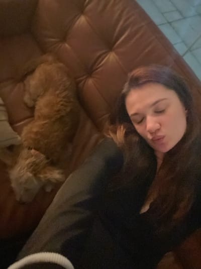 A woman is asleep on a brown leather couch, her face turned towards the viewer. She has long brown hair and is wearing a dark-colored shirt. A small brown dog is curled up near her feet, also asleep. Part of a white object is visible in the lower right corner of the frame. The lighting is dim.
