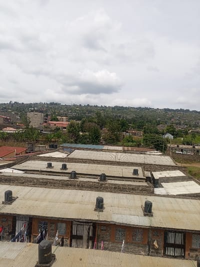An elevated, downward-angled shot depicts a densely populated residential area within Kiamaina ward, Kenya, under an overcast sky. In the foreground and immediate midground, multiple rows of single-story residential structures are visible, characterized by rough-hewn stone lower walls and reddish-orange plastered upper sections. These buildings feature corrugated metal roofs, upon which numerous black plastic water storage tanks are mounted. Rectangular windows are fitted with dark security grilles, and entryways appear as dark, barred doors. Laundry is observed hanging on lines outside several residences in the lower left and center. Beyond these immediate structures, the terrain rises, revealing a continuous expanse of residential buildings, interspersed with green trees and foliage. A multi-story building under construction, featuring exposed concrete framework, is visible in the mid-left. The background consists of rolling hills densely covered with vegetation and numerous distant dwellings, extending to meet a uniform grey, cloudy sky. No individuals or explicit human interactions are discernible.