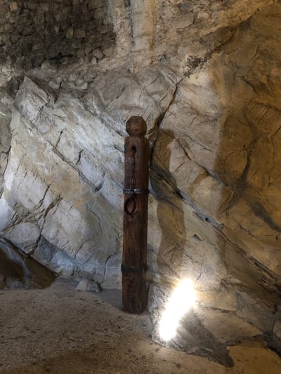 The image shows a dark, cavernous space constructed from rough-hewn stone. The walls are irregular and fractured, with variations in color and texture indicating different types of stone or layers of construction. A wooden beam spans across the space, secured with iron bands. A chain hangs from the beam. The upper right of the image is intensely illuminated, possibly from an artificial light source. The location is likely the Château de Chillon in Veytaux, Switzerland, specifically an interior area possibly used for torture, as indicated by the beam and chain. No people are visible.