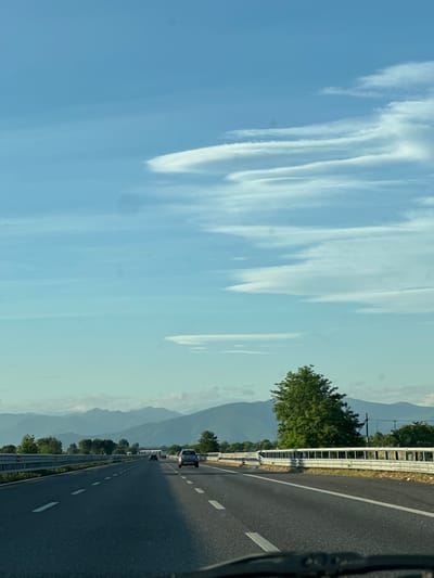 An asphalt highway stretches towards distant mountains under a bright blue sky dotted with wispy white clouds. The highway has two lanes in each direction, separated by a solid white line, and a dashed white line dividing lanes within each direction. Several vehicles are visible on the road, including a silver car in the right lane and a dark-colored car in the left lane. On the right side of the highway, a metallic guardrail runs parallel to the road, with a lush green tree standing tall behind it. The background features a range of hazy, blue-toned mountains. The image is captured from a vehicle traveling on the highway, with the dashboard visible at the bottom. The location is identified as the city of Leno, Italy.