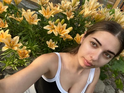 This media file is a selfie taken by a young woman lying down outdoors among a patch of blooming lilies. The scene appears to be a garden or natural area, with rocks and foliage visible in the background. The woman is wearing a black top with white trim and light-colored shorts. Her expression is relaxed and her gaze is directed towards the camera. The lilies are a pale yellow or peachy color with prominent dark spots on their petals. The lighting suggests it is daytime, likely late afternoon or early evening, given the warm, golden tones of the sunlight illuminating the scene. There is no visible text in the image. The overall mood is serene and natural, capturing a moment of quiet enjoyment in a floral setting.