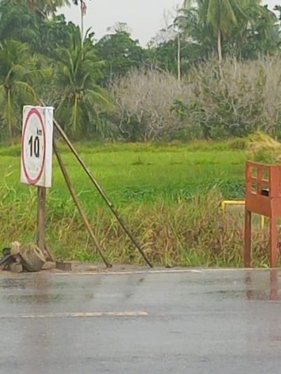This image depicts a roadside scene in Putoh Sa, Indonesia. The focal point is a speed limit sign displaying "10 km". The sign is mounted on a wooden post, which is supported by two angled wooden planks. A pile of rocks and a damaged brick are visible at the base of the sign post. The road surface is wet, indicating it may have been raining. Beyond the road, there is a field of green grass and vegetation, with a line of trees, including palm trees, in the background. To the right of the sign, a rusty metal structure with square openings is partially visible. The sky appears overcast.