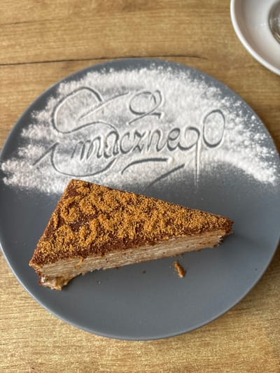 A slice of layered cake with a crumble topping sits on a gray plate dusted with powdered sugar forming the word "Smacznego" (Bon appétit in Polish). The plate is positioned on a wooden surface, likely a table. A white saucer with a sliver of a cup is visible in the upper right corner. The scene is captured in Hajnówka, Poland.