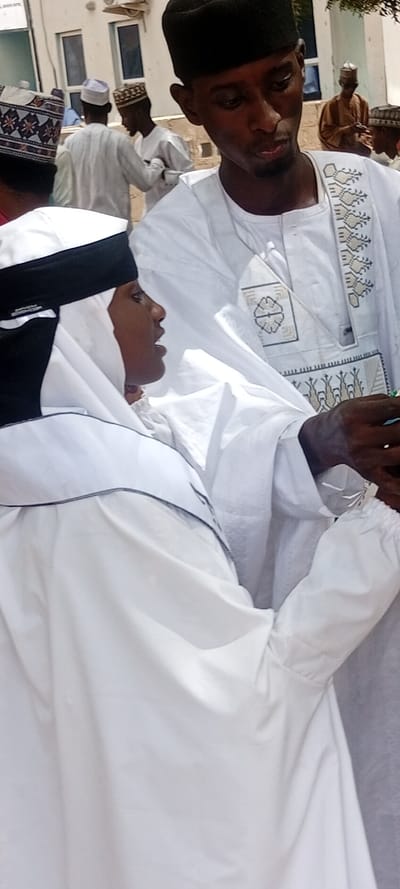 The image displays an outdoor scene in Sani Mai Nagge, Nigeria. In the immediate foreground, two dark-skinned male individuals are prominently featured. The male on the right wears a solid black skullcap and a white traditional tunic-style garment embellished with elaborate tan and white geometric embroidery on the chest and neckline. He looks downwards and slightly to his left. To his left, another male is seen in profile, wearing a white head covering with a distinct black band at the forehead, paired with a white robe featuring a structured collar. His mouth is slightly open, suggesting speech. His right hand is visible in the lower right quadrant, holding a small object. In the midground and background, several other males are present, also dressed in traditional Nigerian attire, including various skullcaps (some patterned, some solid white or tan) and flowing robes. The background features a light-colored building with rectangular windows and an external utility unit mounted high on the wall. The ground appears to be dry, light-colored earth or pavement under daylight conditions.