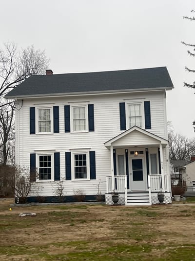 A two-story residential structure, identified by the address number "923" above its main entrance, is depicted under an overcast sky in the Town of Fishkill, United States of America. The house features white horizontal siding and a dark gray asphalt shingle roof with a visible brick chimney on the left side. The front facade presents two double-hung windows on the first floor and two on the second, all framed in white and flanked by dark blue functional shutters. A central white portico with a gable roof shelters the front door, supported by two white square columns. The portico includes a white railing system with vertical pickets and lattice skirting below. Two dark gray urn-style planters are positioned at the base of the portico steps. The front door is white with a small upper window panel, and an exterior light fixture is mounted above it. The immediate foreground consists of a dry, patchy grass lawn with visible dirt. Sparse, leafless deciduous trees frame the house on the left and rear, with a fragment of an evergreen tree visible on the right. A low, dark-colored landscape border runs along the base of the house, with minimal shrubbery. A small yellow marker flag and a cluster of rocks are visible on the ground in the lower-left portion of the image. No individuals or active subjects are present.