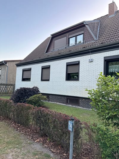 The image shows the exterior of a two-story house at 33 Hauptstraße, Wustermark, Germany. The house has a white brick facade on the lower level and brown shingled siding on the upper dormer. There are several windows with dark frames and brown roller shutters. A dark gray roof with visible tiles covers the house, and a small chimney is visible on the right side. The property features a well-maintained garden with green grass, a hedgerow of dark red and green bushes along the front, and a few trees. A wooden fence borders the property to the left. A metal signpost with a blue and white sign displaying "HA. Wasser 10,9 2,4" is located in the front yard. The sky is clear and light blue, suggesting it is daytime. No people are visible in the image.