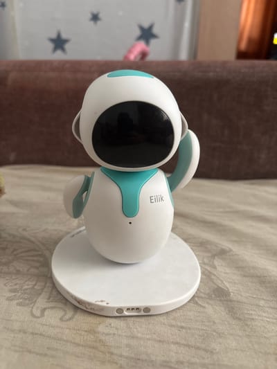 A white and teal robot with a black screen for a face is placed on a white circular charging base. The robot has two small white arms and a teal accent around its neck. The word "Eilik" is printed in grey on its body. The charging base has visible scuff marks and charging contacts on its side. The robot is positioned on a patterned surface that appears to be a tablecloth or bedspread. In the background, a brown upholstered piece of furniture is visible, and a wall with star-shaped decals can be seen above it. The overall environment suggests an indoor setting, possibly a home. The image does not provide any information regarding the location in the city of Клейнікаўскі сельскі Савет, Belarus.