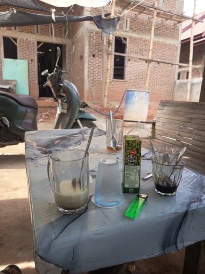 In Rayeukmatang, Indonesia, a scene unfolds on a patterned table. A glass of light brown liquid, likely a beverage, sits in the foreground. Beside it, a plastic cup, possibly for water, contains condensation. A green lighter rests on the table, alongside a carton of "ABC Sari Kacang Hijau" (ABC Green Bean Drink) with a straw. A second glass holds a dark liquid, possibly coffee, with a spoon inside. Another glass, partially obscured, contains a small amount of dark liquid. In the background, a green and silver scooter is parked. Beyond the scooter, a brick building under construction is visible, with scaffolding and roofing materials. The ground is sandy, and shadows suggest it is daytime.