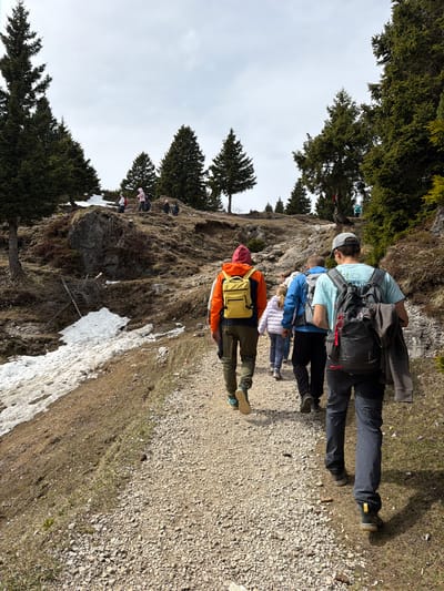 The image shows a group of hikers on a trail in a mountainous region. The location is likely near Kamnik, Slovenia, based on the mountainous terrain and vegetation. A family of hikers are ascending a mountain trail in the spring. The adults have backpacks. There are trees and snow patches along the trail. The sky is overcast. The hiking trail is composed of small stones.