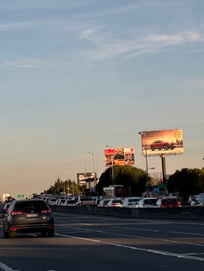 Image depicts heavy traffic on a multi-lane highway in Munro, Argentina. Numerous cars, mostly white, are bumper-to-bumper in at least three lanes. A dark-colored car with license plate "AG 567 IR" is prominently visible in the foreground. Several large billboards are positioned along the highway, advertising Nissan Kicks and McAlister. Overhead signs are present. The sky is light blue with wispy clouds. Street lights line the road. Trees are visible intermittently along the side of the road.