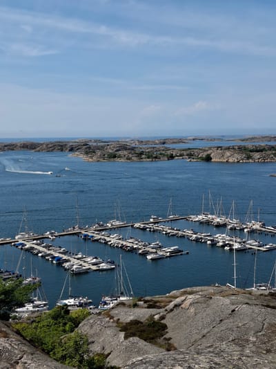 This aerial photograph captures a scenic view of the archipelago in Fjällbacka, Sweden. The foreground is dominated by rocky terrain covered with sparse vegetation, leading down to a tranquil harbor. The harbor itself is filled with a multitude of boats, primarily white sailboats and smaller motorboats, neatly docked. These vessels are moored along wooden piers that extend into the deep blue water.
Beyond the harbor, the expansive sea stretches out, dotted with numerous small, rocky islands. These islands, characteristic of the Swedish coast, are largely unpopulated and covered with low-lying shrubs and trees. The sky above is a clear, bright blue with scattered, wispy clouds, suggesting a pleasant day. In the distance, a few boats can be seen actively navigating the waters, leaving white wakes behind them. The overall scene evokes a sense of peaceful maritime activity and the rugged beauty of the Swedish coastline.