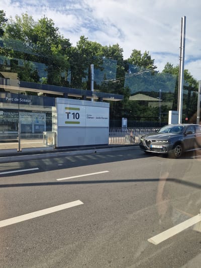 The image depicts the T10 tram stop in Antony, France, at the Parc de Sceaux station. A dark gray Alfa Romeo Tonale is seen driving past the tram stop. The tram stop itself features a modern, glass-enclosed structure with clear signage indicating "Parc de Sceaux" and "T10 Direction Clamart - Jardin Parisien." Reflections of trees and the sky are visible on the glass surfaces. A person in dark clothing can be seen in the background near the tram stop entrance. The road in the foreground has white lane markings. The lighting suggests it is a sunny day with some clouds in the sky.