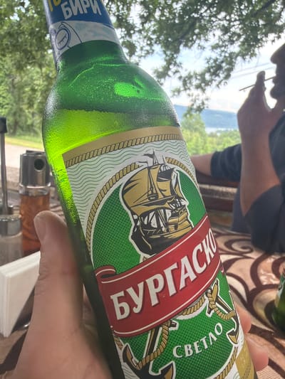 This image depicts a casual outdoor scene in Sushina, Bulgaria, under bright daylight. In the foreground, a hand holds a cold, green glass bottle of beer, evidenced by the condensation on its surface.

The bottle's label is prominent, featuring Cyrillic script. A red band displays "БУРГАСКО" (Burgasko), identifying the brand. Below this, on a green background, the word "СВЕТЛО" (Svetlo), meaning "light," indicates the beer type. The label also showcases a detailed illustration of a sailing ship, surrounded by anchors and rope patterns. On the bottle's neck, "БИРИ" (Biri), plural for beer, is visible.

In the upper left background, a person is partially visible, seen from the chest up, seemingly relaxed. They are holding a thin object, possibly a cigarette or a straw, to their mouth. The backdrop is filled with lush green trees and foliage, suggesting a natural and verdant environment. Beyond the immediate trees, a hazy, distant landscape, likely mountains or hills, is visible under a bright sky, reinforcing the outdoor setting. The presence of a patterned tablecloth under the bottle hints at a table, suggesting a relaxed gathering on a patio, balcony, or outdoor eating area. The overall mood is calm and informal.