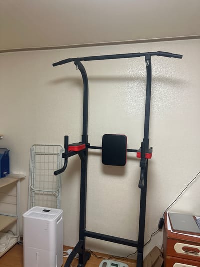 The image displays an indoor scene featuring a black exercise power tower with red and black padded components, centrally positioned. The power tower includes a pull-up bar at the top, dip handles, and back/arm pads. The walls are covered in light beige wallpaper with a subtle, repeating textured pattern. The ceiling is a similar light beige. The floor appears to be light wood.
To the left of the power tower, a light wooden open shelving unit is partially visible, with a blue container on its top shelf. A folded white metal clothes drying rack stands behind a white rectangular dehumidifier, which has a black display panel on its top surface. An electrical cord from the dehumidifier extends towards the floor.
To the right of the power tower, a wooden dresser or cabinet with two drawers is visible; the top drawer is orange-brown with a light cream accent and a metal handle, and the lower drawer is light cream with a similar handle. A silver laptop rests on top of the dresser. A black power adapter and cable are connected behind the dresser, with a thin black wire running vertically along the wall. Partially visible on the floor near the right base of the power tower are a pair of paper bags and a small, light-colored digital scale. No individuals are present in the scene.