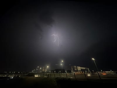 This image captures a nighttime scene of a highway in the vicinity of Badhoevedorp, Netherlands, during a thunderstorm. A fork of lightning illuminates the dark, cloudy sky in the upper center of the frame. The foreground shows a roadside with a fence and some vegetation. Beyond the fence, a highway stretches into the distance, lined with streetlights. Several large overhead gantries, typical of highway signage, are visible. The overall impression is a dramatic and somewhat ominous view of the natural phenomenon over a man-made infrastructure. There are no people or vehicles clearly discernible in the image.