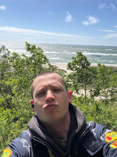 The image shows a person taking a selfie in front of a body of water with a beach and trees. The person is wearing a dark jacket with patches on the sleeve and appears to have an earbud in their ear. The sky is blue with some clouds. The location is identified as Povorovo, Russia.