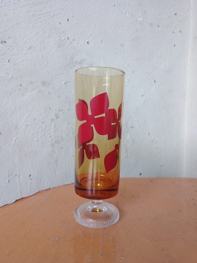 A tall, amber-colored glass with a clear, fluted base stands on an orange-brown surface against a white, textured wall. Red floral patterns are visible on the glass. The scene appears to be indoors, with no other objects or people present. There is no indication of the location being in Jonggol, Indonesia.