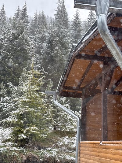 The image captures a winter scene, looking outwards from beneath a wooden structure during a snowfall. The foreground is dominated by the underside of a wooden roof, constructed with light brown planks and darker support beams, showcasing rustic carpentry. A shiny, galvanized metal gutter system runs along the eaves, collecting falling snow and meltwater, curving into a downspout on the left.
Numerous individual snowflakes are visible in the air, creating a soft, bokeh-like effect against the darker wooden elements and the lighter background.
Beyond the wooden structure, a dense forest of evergreen trees is covered in a fresh blanket of snow. The branches of the coniferous trees, likely firs or spruces, are heavily laden with white snow, highlighting their intricate structures. The overall atmosphere suggests heavy, continuous snowfall, with the background trees fading into a misty, overcast sky, characteristic of a snowy winter day.
There is no visible human activity or discernible text in the image. The setting is clearly outdoors, in a natural, forested environment, likely in a mountainous or rural area like Pobit Kamak, Bulgaria, during daylight hours, although the heavy snow clouds obscure direct sunlight. The scene evokes a sense of cold, quiet winter serenity.