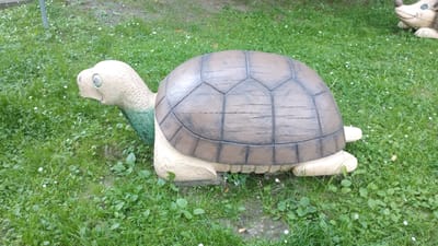 A concrete statue of a turtle is situated on a grassy lawn. The turtle is depicted with a tan-colored head and legs, and a dark brown shell with lighter brown lines delineating the scutes. Its eyes are large and black. The neck of the turtle is a faded green. The statue is positioned in the center of the frame, facing left. The background consists of a green lawn dotted with small white flowers. To the right of the turtle, a portion of another statue, appearing to be a hedgehog, is visible. The lighting suggests it is daytime, and the weather appears to be mild, possibly overcast. There is no visible text in the image. The scene is likely an outdoor decoration in a public space or garden in Khmilnyk, Ukraine.