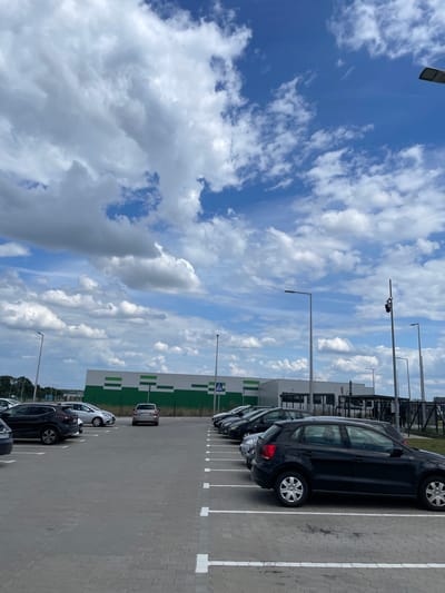 This is an outdoor, eye-level, medium shot of a parking lot in Nowa Wieś Kącka, Poland. The sky is filled with large, white, fluffy clouds against a bright blue backdrop. In the foreground, a black Volkswagen Polo is parked. To the left, other cars are visible, including a silver Skoda Fabia. In the background, a large building with a green and white striped facade is situated behind a row of parked cars. Several streetlights are positioned throughout the parking lot.