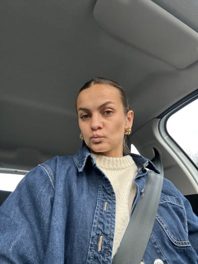 A light-skinned adult female is depicted seated within a vehicle, identified as being in Saint-Cyr-sur-le-Rhône, France. She is positioned to face the viewer directly, exhibiting a slight pout with her lips. Her light brown hair is pulled back from her face. She wears a cream-colored knitted sweater layered under a dark blue denim shirt or jacket featuring visible light-colored stitching and buttons. Ornate, gold-toned stud earrings with a textured design are present on both earlobes. A black seatbelt is securely fastened across her chest and right shoulder. The vehicle's interior consists of a light grey ceiling and a sun visor partially visible above her. A car window on the right side of the frame reveals an overcast sky and indistinct outdoor foliage, possibly bare trees or bushes.