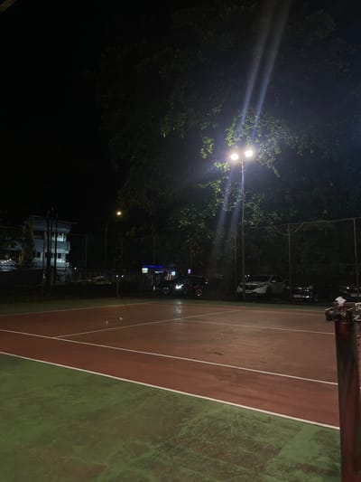 This image depicts an outdoor tennis court at night, illuminated by artificial lighting. The court itself is divided into two main colors: a reddish-brown surface for play, bordered by a green out-of-bounds area. White lines demarcate the playing boundaries. In the background, a chain-link fence encloses the court, behind which several parked cars are visible. A building, appearing to be a multi-story structure with some windows illuminated, stands to the left. Trees with dense foliage are present, with one large tree partially obscuring a light pole with two bright lamps. The sky is dark, indicating nighttime. The overall impression is of a sports facility operational after dark. This scene is located in Manado, Indonesia.