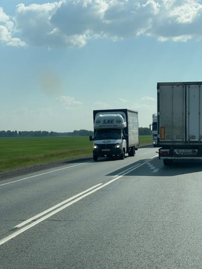 The image depicts a view from a vehicle traveling on a paved road in Chik, Russia, during daytime. The sky is a clear blue with scattered white clouds, suggesting fair weather. The road has visible white lane markings.
The primary subjects are two large trucks. In the foreground, a white truck with a black cab is visible. The side of its trailer displays text, including "S.A.V." and "АВТОАГЕНТСТВО". A license plate is visible on the front of the cab, reading "H930PY 102". Behind this truck, a larger, silver-colored truck with a white trailer is seen. The rear of this truck has visible license plates, one reading "Aa 9705 42", and "SCHMITZ" branding. A smaller white truck is visible between the two larger trucks.
The scene is set along a highway with a strip of green grass and a line of trees visible on the left side of the road, indicating a rural or semi-rural landscape. The orientation of the image suggests it was taken from a car traveling behind or alongside the trucks. There are no people clearly visible in the image. The overall impression is one of a journey on a highway.