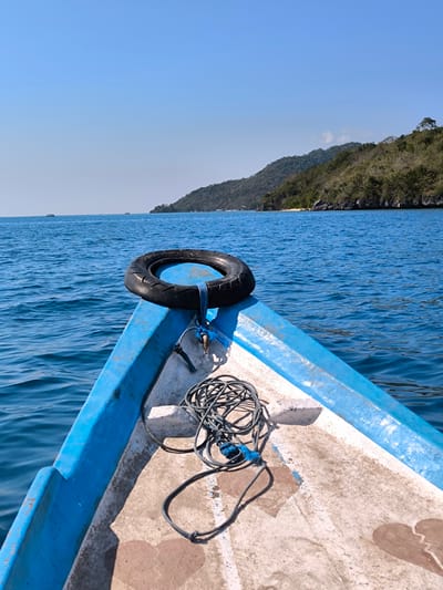 A frontal view of a blue and white boat navigates calm, blue waters under a clear sky. The bow of the boat is visible, with a black tire secured by a blue rope at its tip. A coiled rope lies on the deck near the tire. In the distance, a lush green island with a sandy beach is visible, dotted with trees and some structures. The sea is a vibrant blue, with gentle ripples on the surface. The scene appears serene, suggesting a journey through a tropical coastal area. The location is identified as Tanjung Boleng, Indonesia.