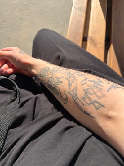 In Priovrazhnoe, Russia, a person sits on a wooden bench outdoors. Their left arm, adorned with tattoos, rests on their lap. The individual is wearing dark clothing, and the scene is illuminated by bright sunlight, casting shadows on the bench. The ground appears to be a light-colored gravel or dirt path.
