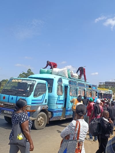 A blue "BENJO Kenya" bus is crowded with people and luggage. Two men in red outfits are on the roof, loading or adjusting bags. Passengers stand near the open bus door and others are further down the road. The bus is parked on a street under a sunny sky with some clouds. A woman carrying a black bag stands in the foreground, watching the activity.