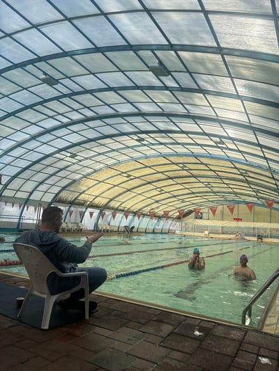 The scene depicts an indoor swimming pool facility in Rehovot, Israel. The pool is covered by a large, arched, translucent roof composed of a grid of metal frames and panels, displaying variations in color and transparency. The facility contains a multi-lane swimming pool with lane lines segmenting the water. Orange pennant banners, some bearing the "Speedo" logo, are suspended above the pool along the far wall. A red digital clock on the far wall displays "10:04." Stacks of white plastic chairs and a lifebuoy are also visible along the far wall.
In the foreground, a male individual is seated in a white plastic chair on a dark mat beside the pool. He has short dark hair and a beard, and is wearing a grey hooded sweatshirt and grey sweatpants. He is facing the pool, gesturing with an open right hand towards the swimmers. A dark cup is situated on the mat near his feet.
Within the mid-ground of the pool, two adult males are partially submerged. The closer individual, with a mustache, wears a blue swimming cap and clear goggles, with his hands near his face. The second individual, also with a mustache, wears a purple swimming cap and purple goggles, looking towards the foreground. Additional individuals are visible swimming or standing in the background lanes of the pool. The pool deck consists of reddish-brown pavers, and metal handrails for pool entry are present on the right.