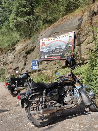 A medium shot captures two motorcycles parked on a dirt road, with a rocky incline and lush green foliage forming the backdrop. The motorcycle in the foreground, a black Royal Enfield Bullet, is angled slightly to the right, showcasing its engine, exhaust pipe, and rear wheel. A black helmet rests on its handlebars. Behind it, a grey motorcycle is parked facing left, its rear visible. A blue sign with white lettering reads "CURVE NO. 8 RD 2120". A large poster is affixed to the rock face above the motorcycles, featuring images of Buddhist figures and text in English and Hindi, promoting the "Buddhist Cultural Society of Key Gompa". The scene appears to be in Siyal, India, given the presence of the "Key Gompa" mentioned on the poster, which is a well-known monastery in Spiti Valley, Himachal Pradesh, India. The lighting suggests it is daytime.