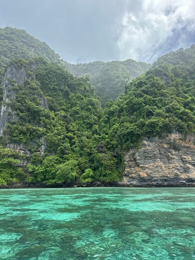 A wide shot captures a dramatic seascape with lush green mountains meeting crystal-clear turquoise waters. The mountains, covered in dense vegetation, rise steeply from the water's edge, with rocky cliffs visible in some sections. The water in the foreground is calm, with sunlight illuminating the underwater topography, revealing coral formations and sandy patches. The sky above is overcast with scattered clouds, lending a soft, diffused light to the scene. The image appears to be taken from a boat, with the camera looking towards the shore. The location is identified as Haad Hin Khom, Thailand. There are no people or visible human activity in the image.