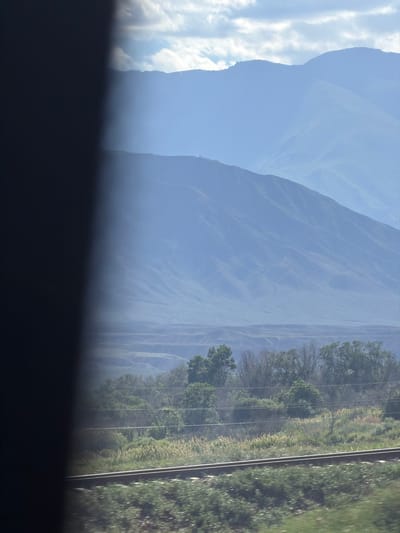 The image captures a distant mountain range veiled in a soft, blue haze. The mountains recede into layers, with the closest range showing more distinct contours and vegetation, while the further mountains are mere silhouettes against a pale sky. In the foreground, a blurry section of dark, vertical material, possibly a window frame or a structural element, partially obscures the view on the left. To the right of this dark section, a landscape unfolds. Tracks of a railway line run horizontally across the lower portion of the image, with blurred green foliage in front of them. Beyond the tracks, a band of trees and shrubbery creates a textured middle ground. The overall scene evokes a sense of travel and observation, likely from a moving vehicle. The location of Kok Moynok Eki, Kyrgyzstan, is a rural or mountainous area, consistent with the depicted scenery of rugged terrain and open skies.