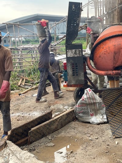 This image depicts a construction site in Okinano, Nigeria. In the foreground, several construction workers are engaged in their tasks. One worker, wearing a brown shirt and blue pants, stands to the left with red gloves on his hands. In the center, another worker, dressed in a brown shirt and dark pants, balances a bucket of what appears to be cement or building material on his head, also wearing red gloves. Beside him, a cement mixer, predominantly orange, is visible, with a portion of its mechanism and hopper in view. To the right, a third worker, in a yellow sleeveless shirt, is seated. In the background, scaffolding made of bamboo supports a larger structure, likely a building under construction, with a modern, angular roofline visible in the distance. The ground is uneven and appears to be dirt and rubble, with puddles of muddy water scattered throughout. A large bag with text, potentially of building materials, is also present near the cement mixer. The overall scene conveys an active construction environment under an overcast sky.