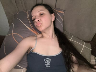 A woman is lying on a pillow, taking a selfie. She has dark hair and is wearing a dark blue tank top with the words "ST. GEORGE LONDON" printed on it. The pillow has an orange and white striped pattern. The background appears to be a bed with a grey blanket. The woman is puckering her lips, and her eyes are open. The scene is indoors, likely a bedroom. There is no indication of the city of Postoyata, Ukraine.