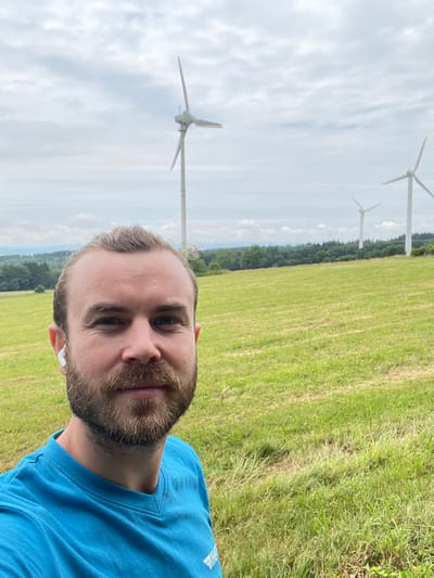 A selfie is taken by a man with a beard and blonde hair pulled back, wearing a blue t-shirt and white earbuds. He is standing in a grassy field with a row of wind turbines in the background, and a wooded area further in the distance. The sky is overcast with some patches of blue. The image appears to be taken in Schwarzenborn, Germany, based on context provided.