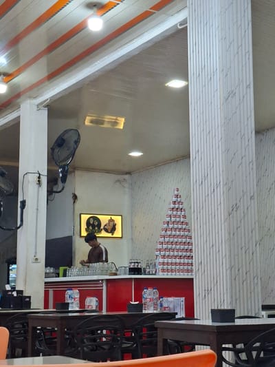 The image shows the interior of a cafe or restaurant in Sigli, Indonesia. In the foreground, there are several dark wooden tables with black chairs, suggesting a dining area. On one table, there are two plastic bottles of water. In the midground, a red counter is visible, with a young man in a brown shirt working behind it, surrounded by stacks of glasses and bottles. To the right of the counter, a tall, pyramid-like structure made of stacked cans is displayed. The walls of the establishment are decorated with a ribbed, textured pattern, and there are ceiling fans mounted on the white columns. A framed sign with cartoonish figures is also visible on the wall. The overall atmosphere appears to be casual and functional.