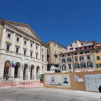 The image depicts an urban scene in Piazza Cavour, Ancona, Italy, under a clear blue sky.

On the left, the neoclassical facade of the Teatro delle Muse stands prominently, characterized by a multi-story structure with a grand portico of tall columns supporting a triangular pediment featuring sculpted relief. At the ground level, several arched openings form an arcade. Three flags are displayed: the Italian national flag (green, white, red), a green and red regional flag, and the European Union flag. Two vertical banners with artistic imagery and text hang from the arches. An orange plastic mesh construction fence encloses the area directly in front of the building, partially obscuring a dark, human-like statue behind it. A bright green utility vehicle is partially visible behind the fencing, near the building's base.

Extending across the mid-ground from the center to the right is a tall, light-colored wooden construction hoarding. Several large posters are affixed to this hoarding, displaying architectural renderings, historical images, and official notices or safety information. Behind the hoarding, a cluster of multi-story residential or commercial buildings rises, featuring varied pastel-colored facades (beige, light pink, yellow) and numerous windows, many equipped with closed green louvered shutters.

The foreground consists of a paved ground surface. A single white and orange striped traffic cone is visible in the bottom-left corner. No individuals are clearly discernible in the scene. The overall impression is one of ongoing urban development or renovation under bright daylight conditions.