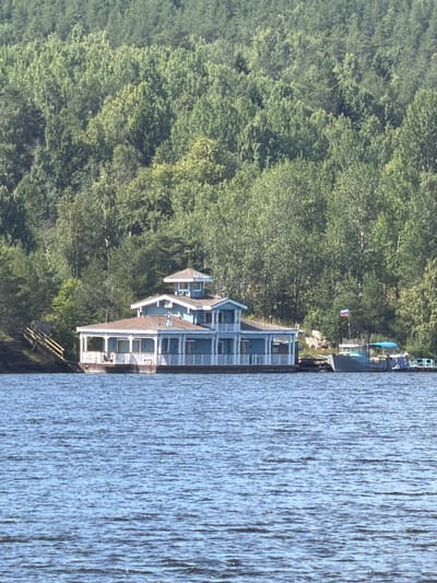The image depicts a light blue, multi-story house situated on the water's edge, backed by a dense forest. The house features a wraparound porch with white pillars and railings, a dark-colored roof, and several windows. A set of wooden stairs with yellow railings leads from the shore to the house. Two boats are docked near the house, one of which displays a Russian flag. The water in front of the house is rippling. The scene appears to be a residential property in Yalguba, Russia, likely on a lake or river, given the presence of boats and the wooded backdrop.