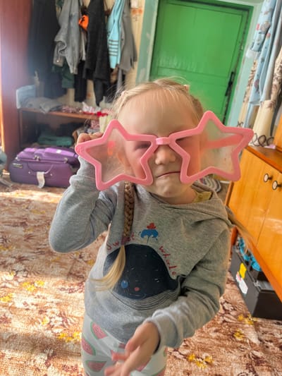 An outdoor scene in Kovriga, Russia shows a young girl in a gray hoodie and patterned pants wearing large pink star-shaped glasses. The girl is standing in a room with a patterned rug on the floor. To the left, there is a purple suitcase and a shelf with clothes. To the right, there is a wooden cabinet with drawers and a green door in the background. The lighting in the room is dim, with sunlight streaming in from the left.